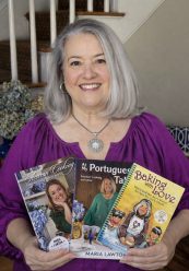 A woman with long gray hair and a purple top smiles indoors, holding Maria's Signed Three Book Bundle. The bundle includes {{brizy_dc_image_alt imageSrc=