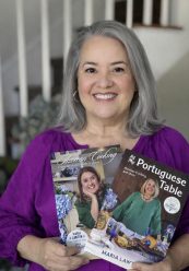 Maria Lawton, smiling with gray hair in a purple top, holds Maria's Signed Two Book Bundle—featuring her photo on both covers. A staircase appears in the background. {{brizy_dc_image_alt imageSrc=