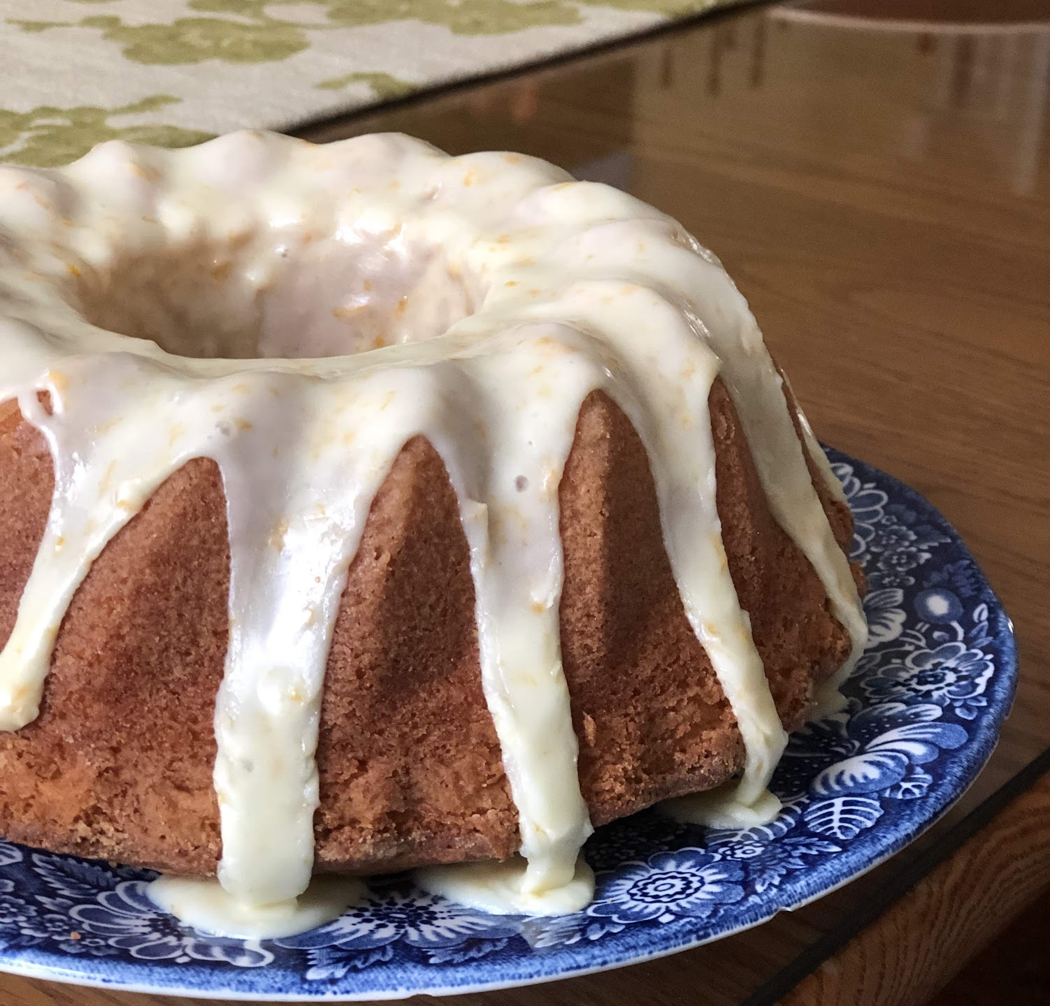 A bundt cake with white icing dripped over the top sits on a blue patterned plate on a wooden table. The cake appears golden brown with visible texture on its sides.