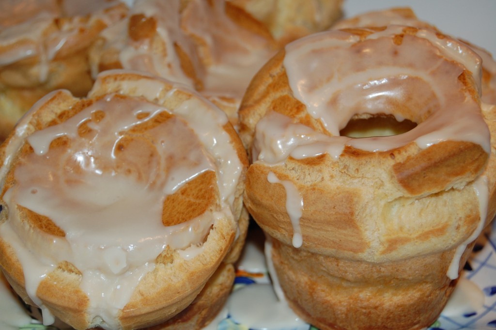 Close-up of a few freshly baked popovers drizzled with a sugary glaze, reminiscent of the delectable pastries from Maria Lawton’s Azorean cooking. The golden brown treats have a puffy, airy texture and are arranged on a plate, with some of the glaze dripping slightly over the sides.