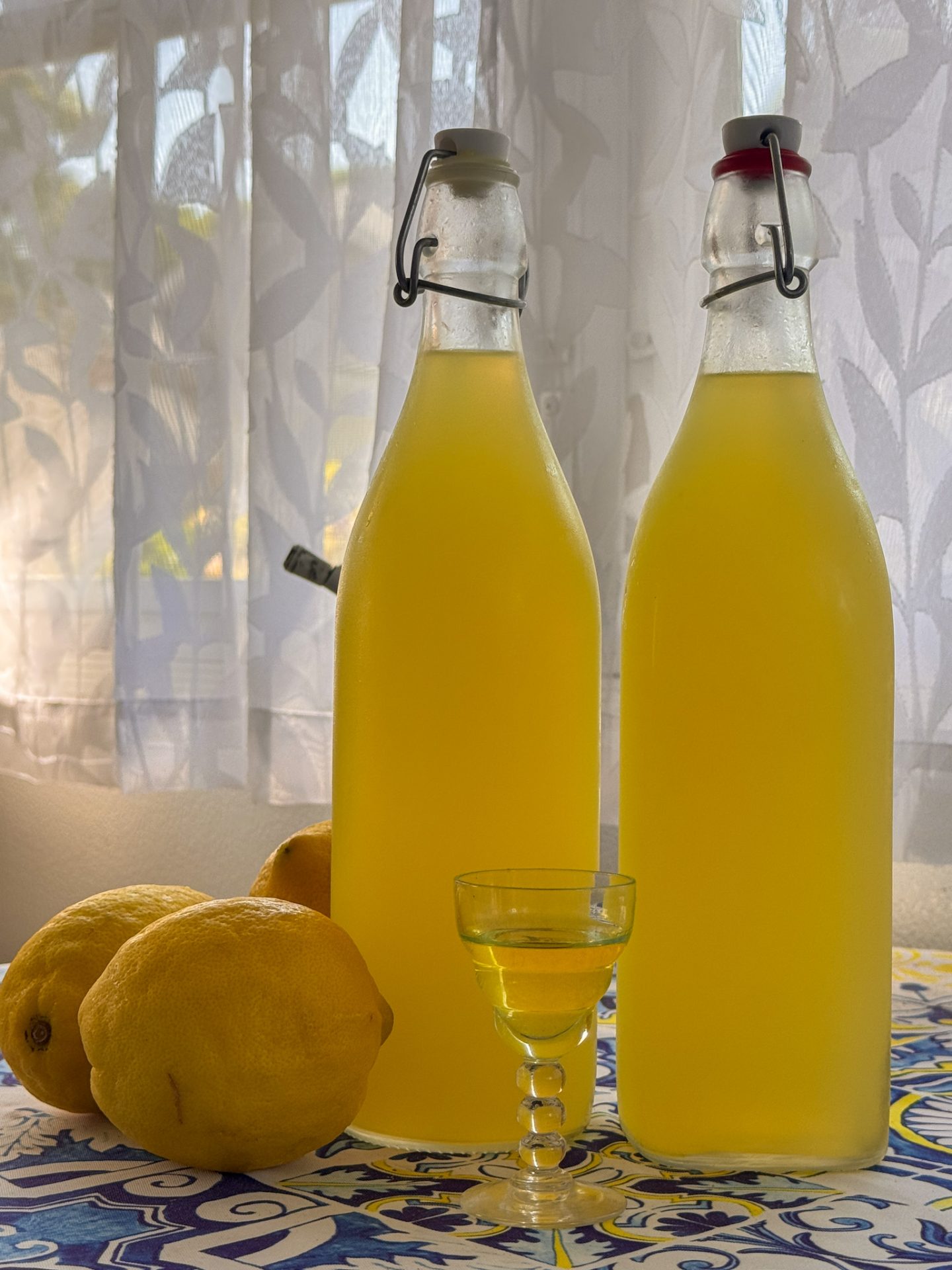 Two glass bottles filled with yellow liquid stand next to a glass of the same, suggesting a homemade Lemon Liqueur recipe. Three whole lemons rest nearby on a patterned surface, as a sheer curtain with a leaf design drapes in the background.