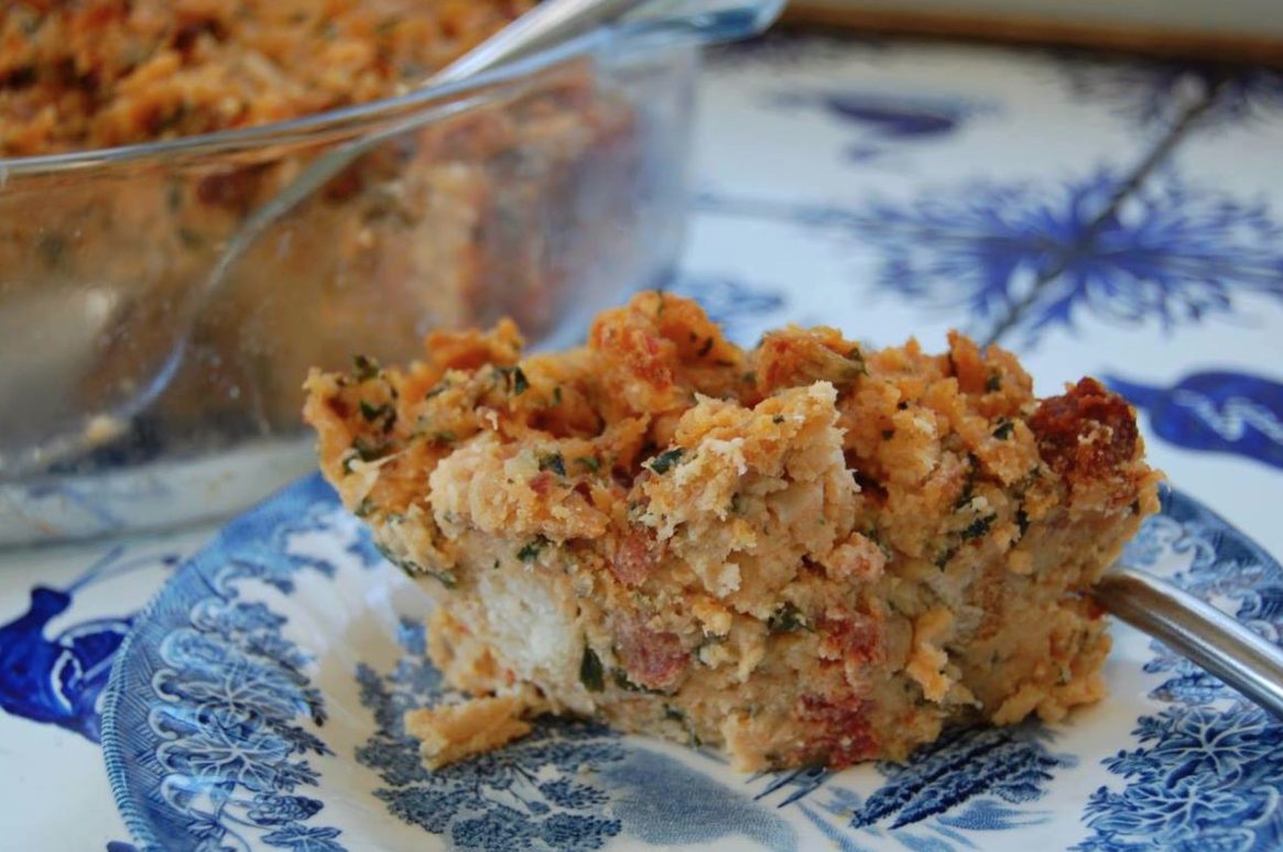 A serving of bread stuffing with herbs is on a blue and white patterned plate, with a glass baking dish containing more stuffing in the background. A metal utensil rests beside the portion on the plate.
