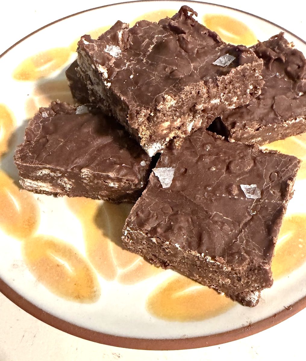 Five chocolate dessert bars are stacked on a yellow patterned plate. The bars contain small chunks of cookies and are topped with a few flakes of sea salt.