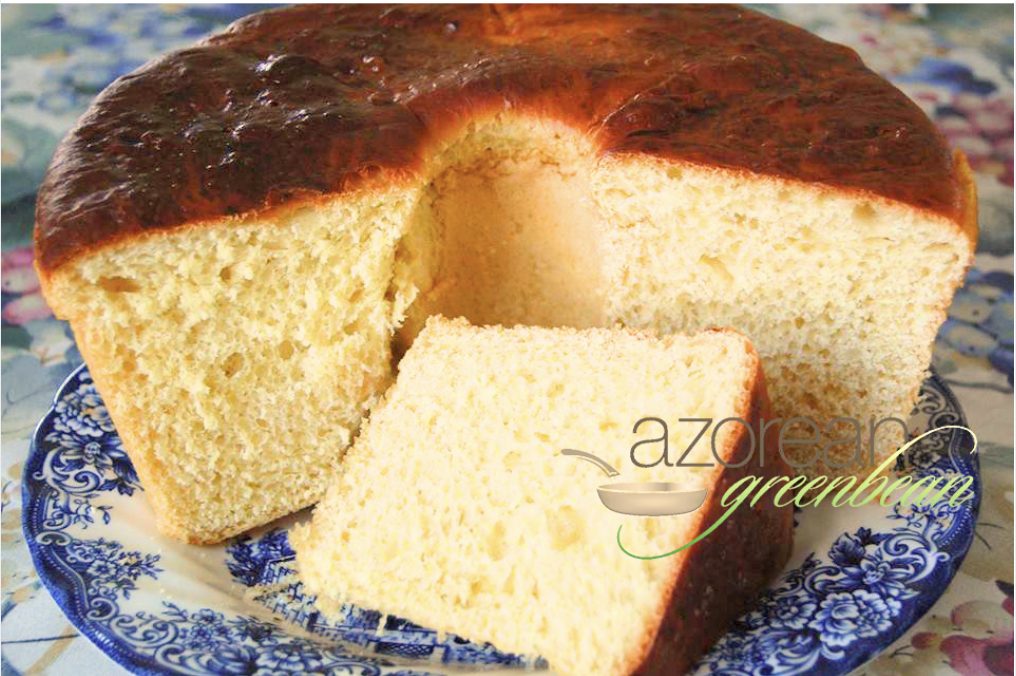 A round loaf of golden-brown sweet bread with a piece sliced out, displayed on a blue and white patterned plate. The bread has a light, airy texture. The words "azorean greenbean" are overlaid in the foreground.