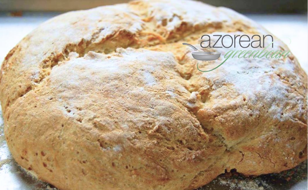 A round loaf of rustic bread with a golden-brown crust sits on a floured surface. The words "azorean greenbean" with a pan icon appear in the top right corner of the image.