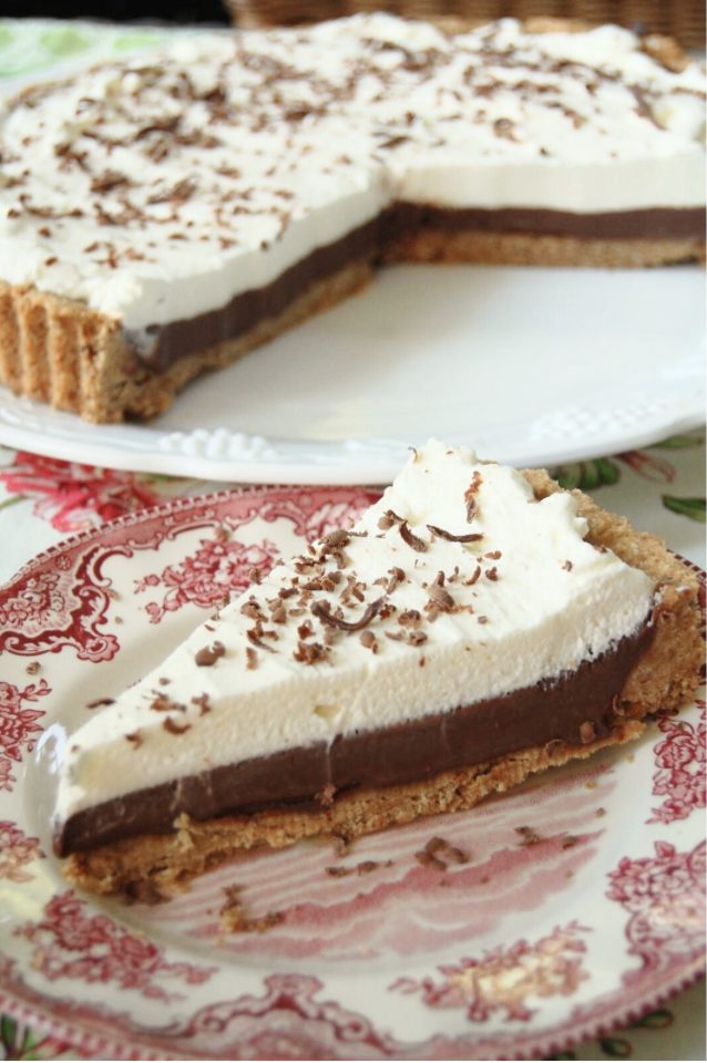 A slice of chocolate cream pie topped with whipped cream and chocolate shavings is served on a decorative plate, with the rest of the pie in the background.