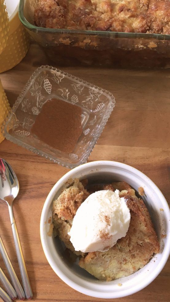 A bowl of bread pudding topped with a scoop of vanilla ice cream and a sprinkle of cinnamon sits on a wooden surface near a clear glass dish of cinnamon, a baking dish of bread pudding, and several spoons.