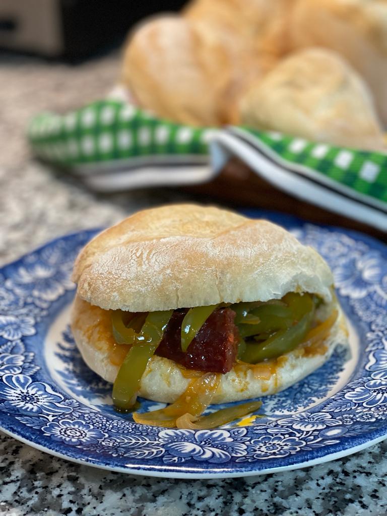 A sandwich with sliced green bell peppers and sausage on a crusty roll is served on a blue and white patterned plate. In the background, more bread rolls and a green-checked cloth are visible.
