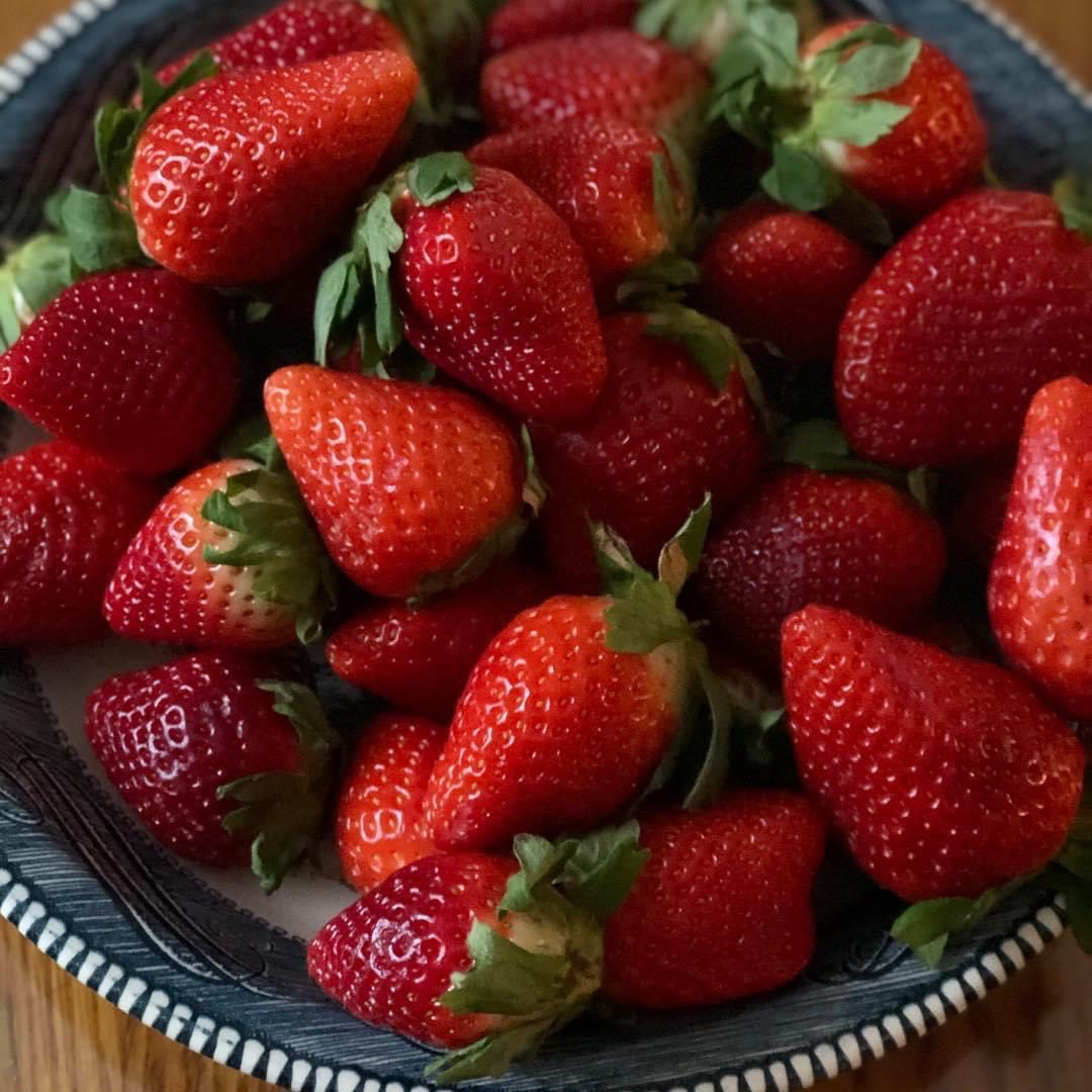 A bowl filled with fresh, ripe strawberries, some with stems attached, sits on a wooden surface. The strawberries are bright red and glossy.
