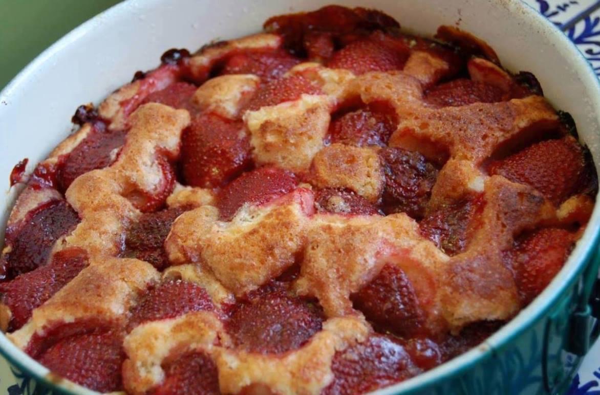 A baked strawberry cobbler in a dish, topped with browned crust and sliced strawberries arranged throughout. The dessert appears freshly baked with edges slightly caramelized.