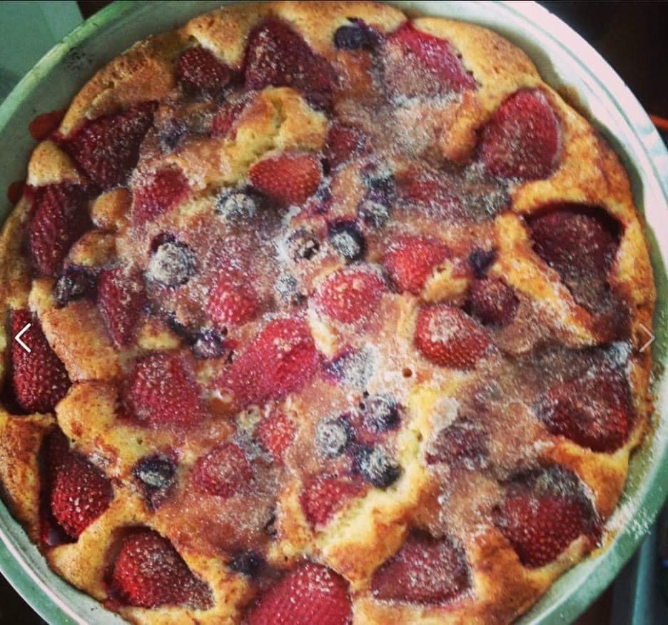 A round cake topped with sliced strawberries and blueberries, sprinkled with sugar, baked in a metal pan.