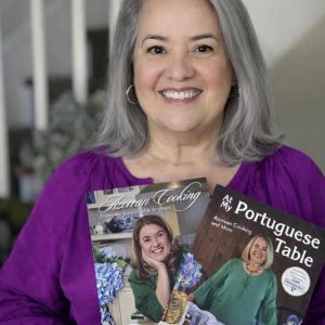 Maria Lawton, smiling with gray hair in a purple top, holds Maria's Signed Two Book Bundle—featuring her photo on both covers. A staircase appears in the background.
