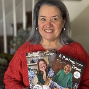 A woman with gray hair and a red sweater stands indoors, smiling and holding Maria's Signed Two Book Bundle. A staircase is visible in the background.