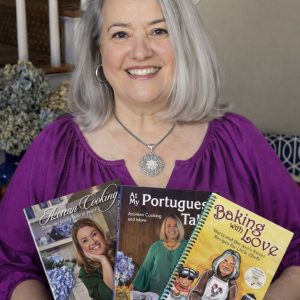 A woman with long gray hair and a purple top smiles indoors, holding Maria's Signed Three Book Bundle. The bundle includes "Azorean Cooking," "My Portuguese Table," and "Baking with Love" by Maria Lawton. A staircase is behind her.