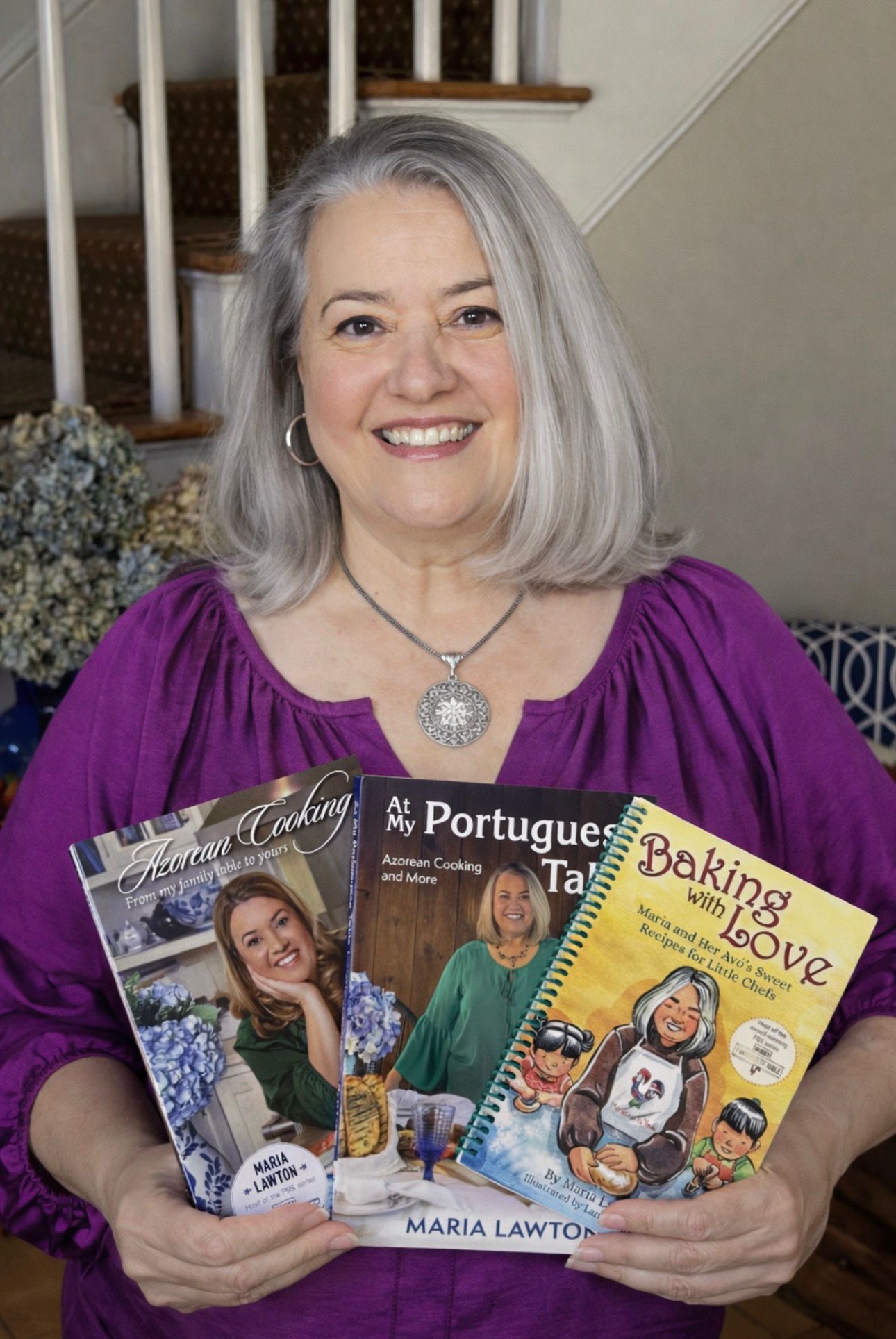 A woman with long gray hair and a purple top smiles indoors, holding Maria's Signed Three Book Bundle. The bundle includes "Azorean Cooking," "My Portuguese Table," and "Baking with Love" by Maria Lawton. A staircase is behind her.