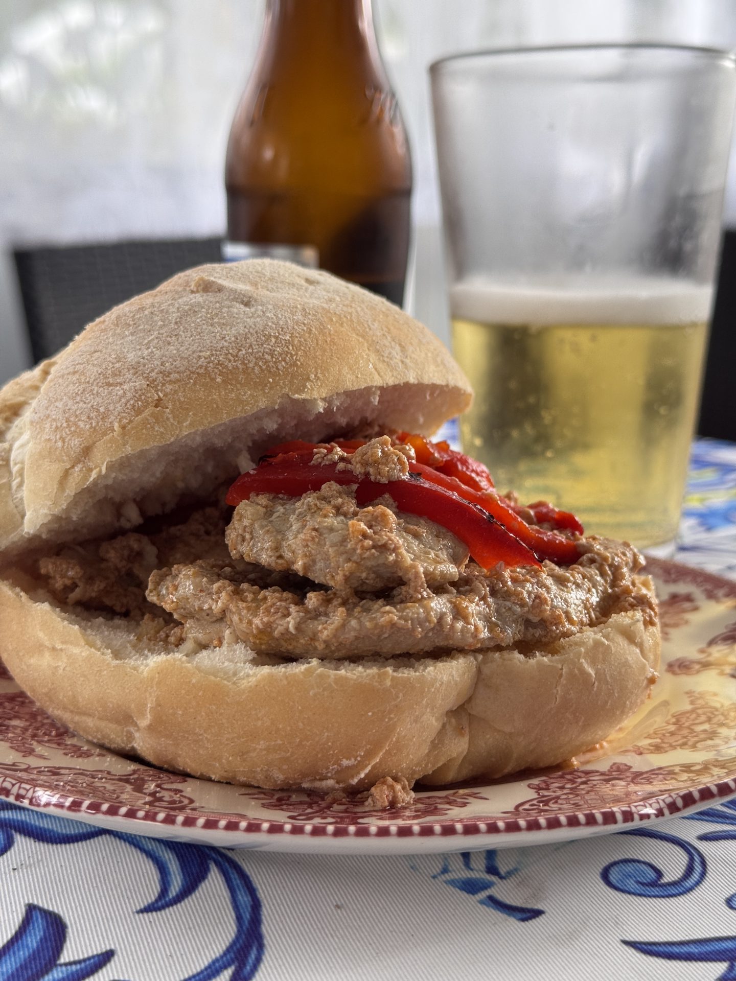 A sandwich filled with a meat spread and red peppers sits on a plate. In the background, there is a glass of beer and a brown beer bottle on a table with a blue and white patterned tablecloth.
